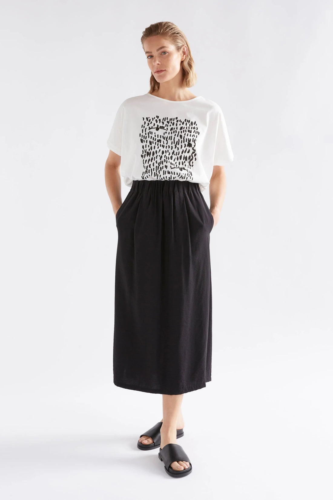 woman standing hand in the pockets wearing black skirt and elk bruk tee with black tropea placement print and black slippers
