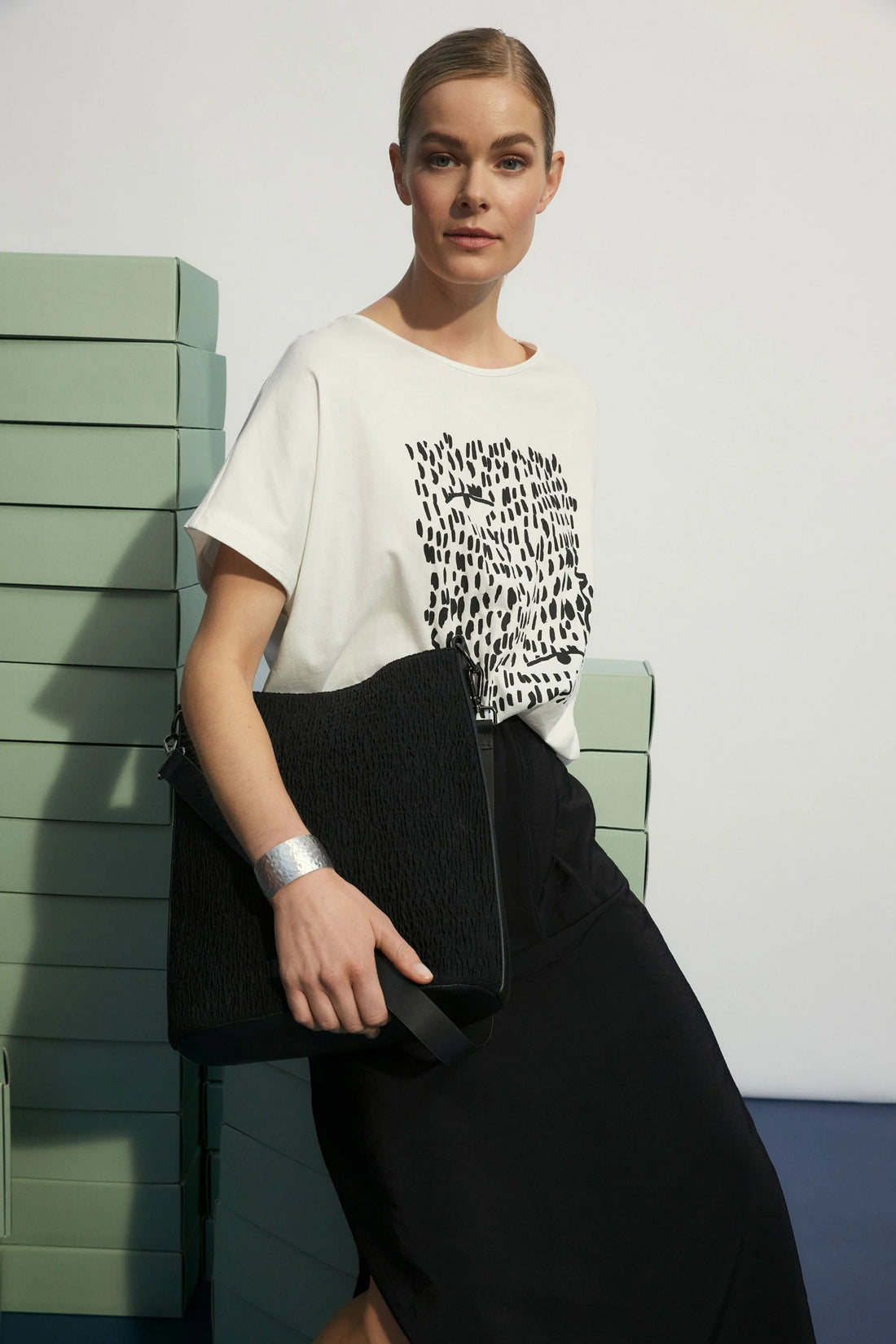 Woman sitting in stack green boxes holding a black bag, wearing  black skirt and elk bruk tee with black tropea placement print