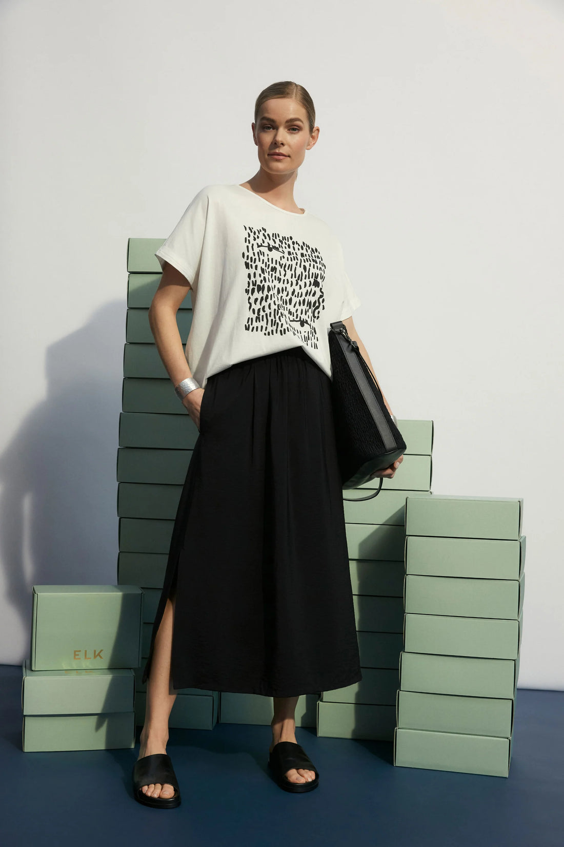 woman standing wearing black skirt and elk bruk tee with black tropea placement print, and black slippers holding black bag