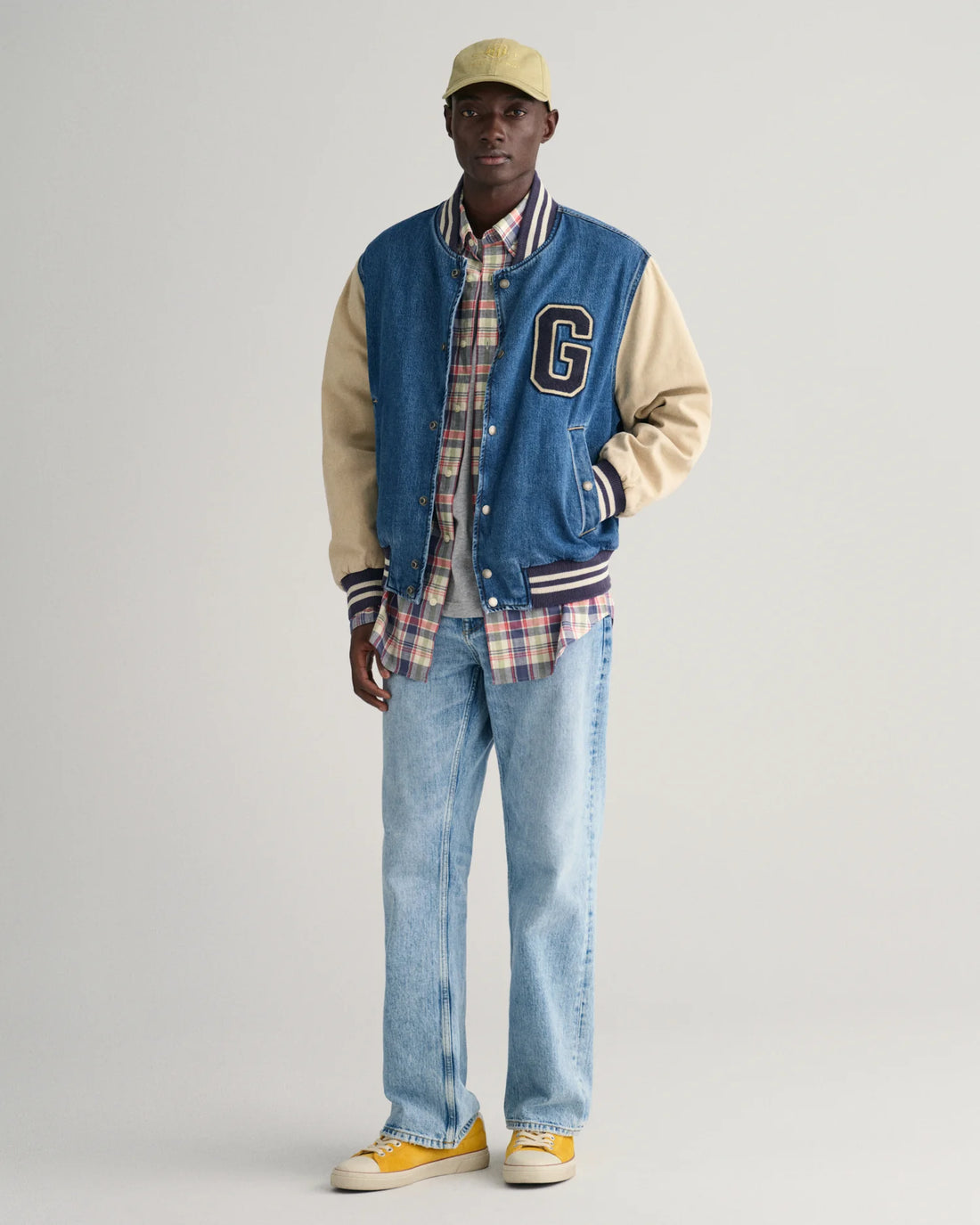 man wearing cap, denim pants, grey shirt, gant regular fit checkered cotton linen shirt in dusty blue sea, and varsity jacket, and yellow shoes