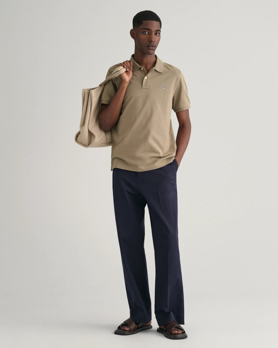 man holding a bag wearing gant regular fit shield pique polo shirt in dried clay, pants, dark brown slip on slippers