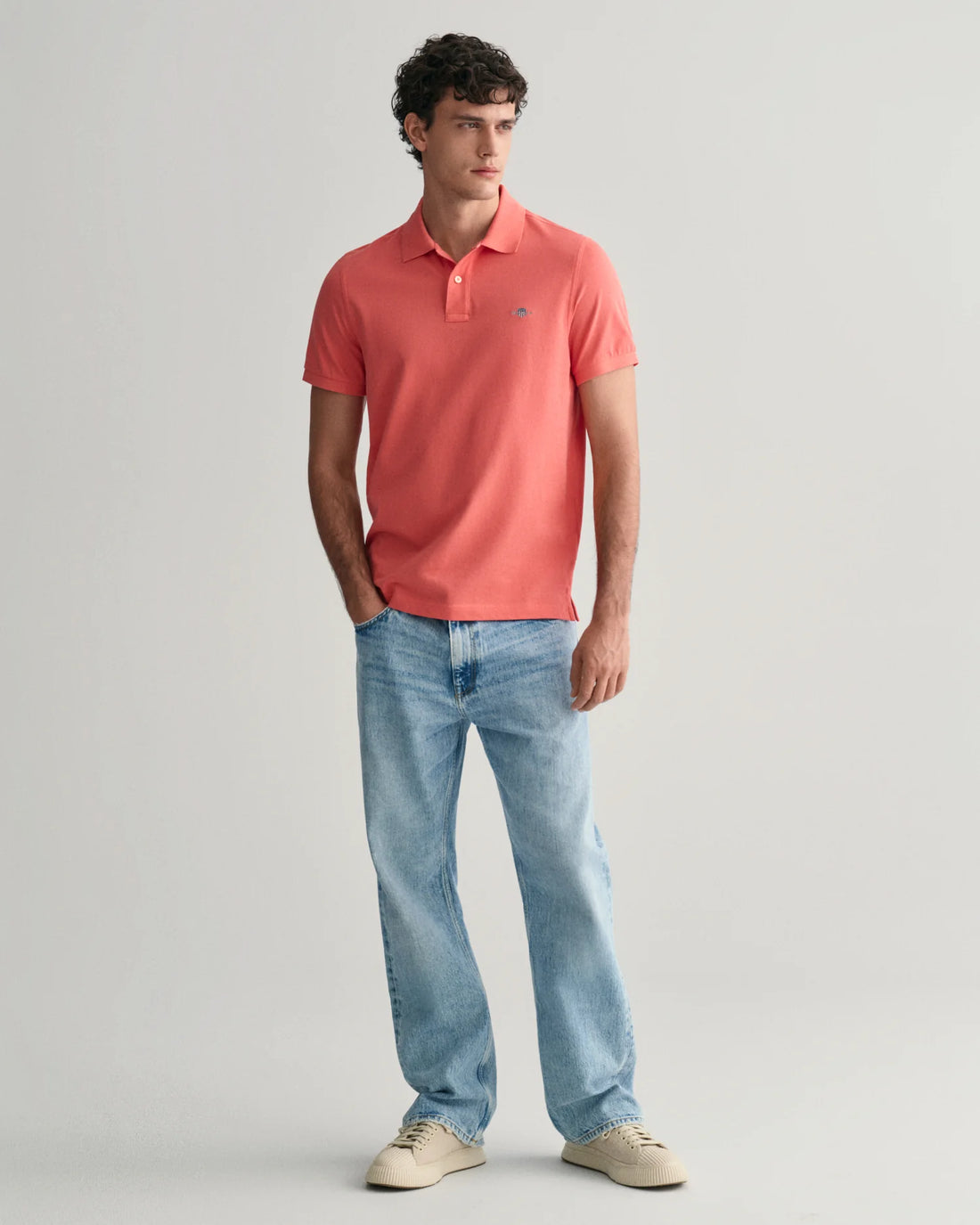 man standing wearing gant regular fit shield pique polo shirt in sunset pink, denim pants, and shoes