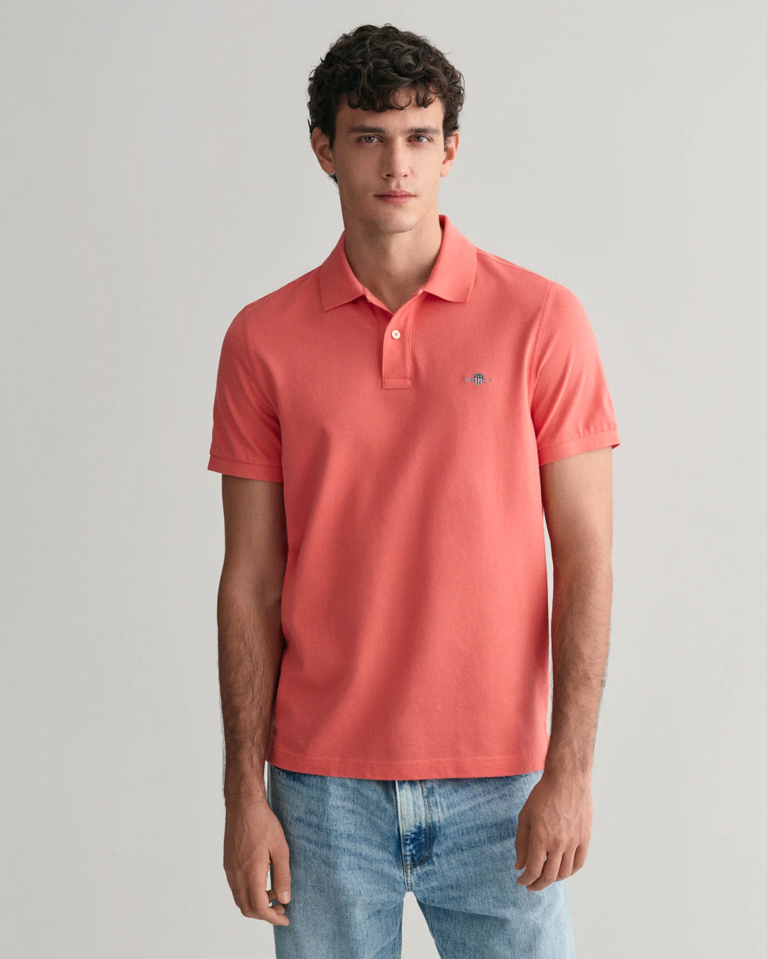 man standing wearing gant regular fit shield pique polo shirt in sunset pink, denim pants