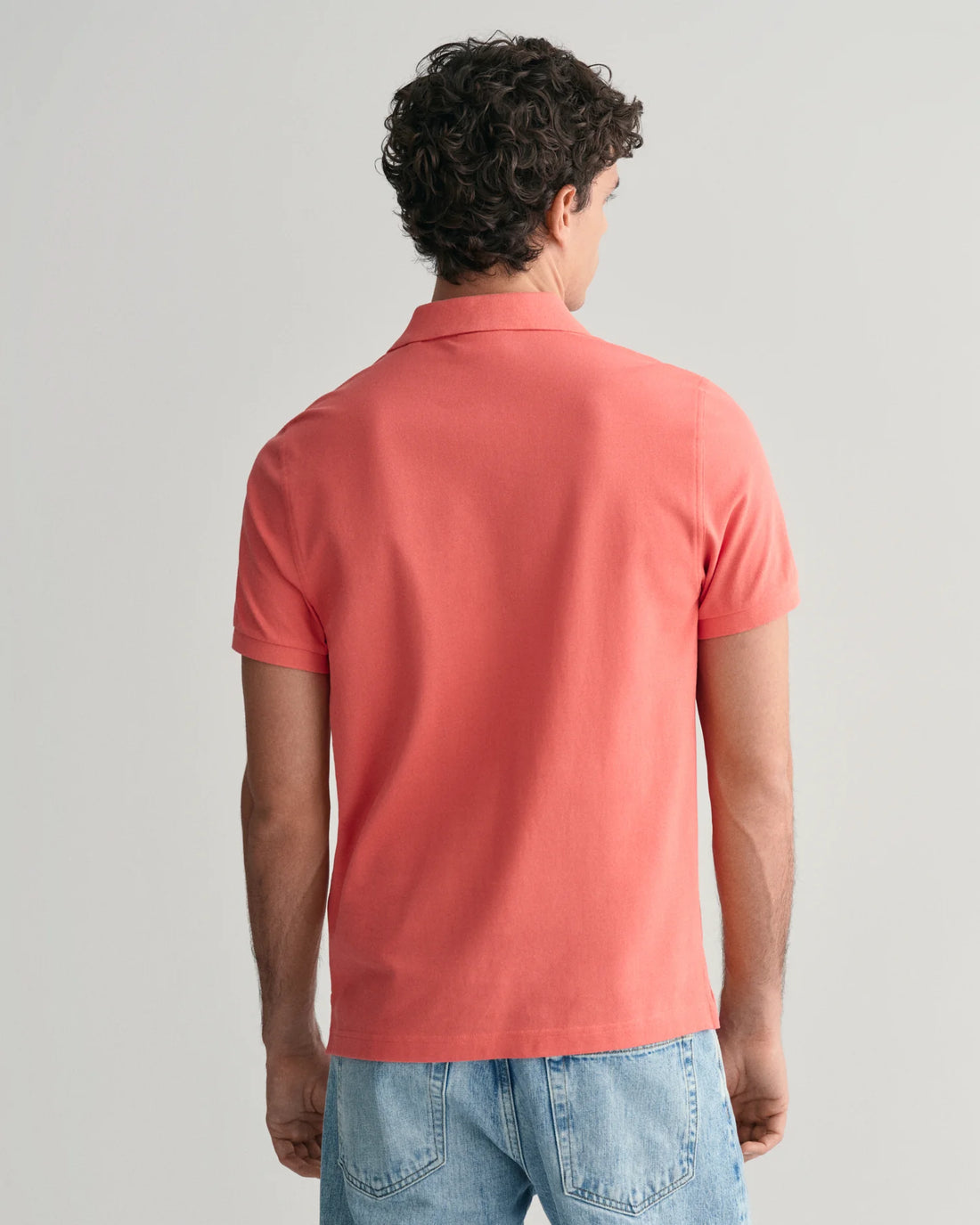 man standing facing the back wearing gant regular fit shield pique polo shirt in sunset pink, denim pants