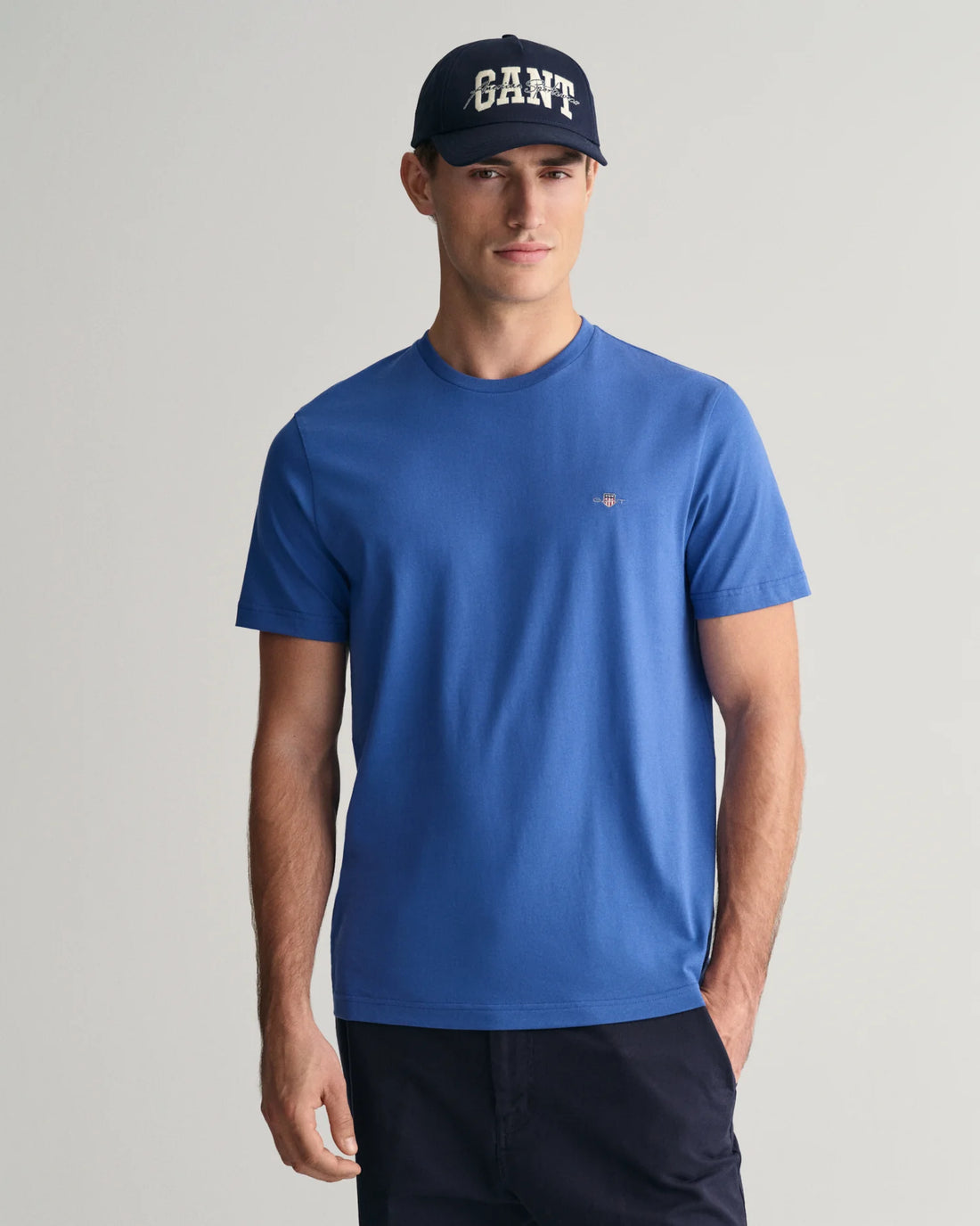 man standing wearing gant shield t-shirt in rich blue, navy blue pants, and navy blue cap