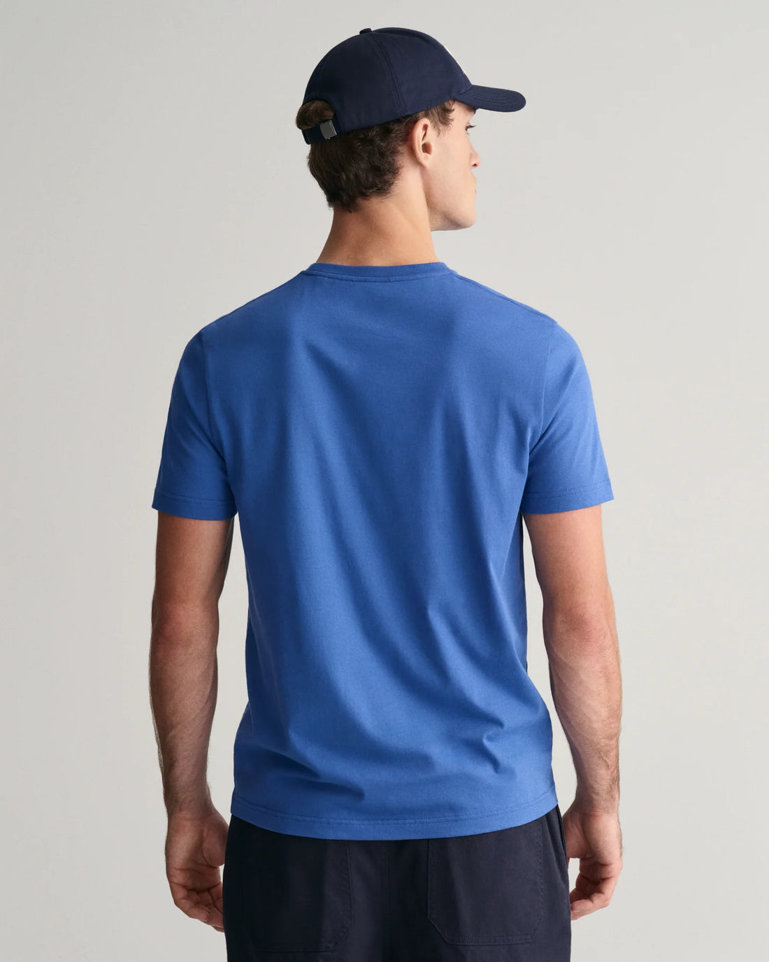 man facing the back wearing gant shield t-shirt in rich blue, navy blue pants and navy blue cap